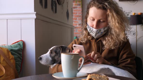 Woman Wearing Face Mask Sitting In Cafe With Dog Using Smartphone