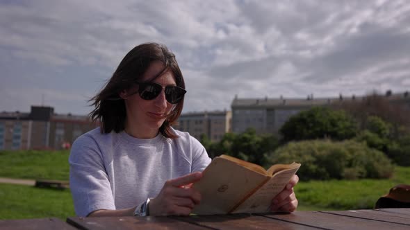 A Girl in the Park Sits at a Wooden Table and Reads a Book