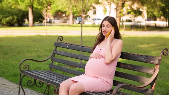 Smiling Pregnant Woman Sitting on Bench in Summer City Park and Talking on Her Mobile Phone