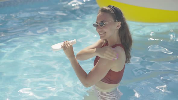 Middle Shot of Smiling Tanned Girl in Sunglasses Standing in Pool Outdoors and Applying Suntan Cream