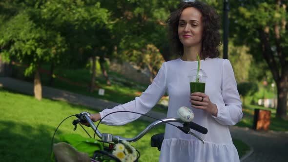 Beautiful Woman in a White Shirt and Blue Jeans Walking in the City Park Holding Her City Bicycle's