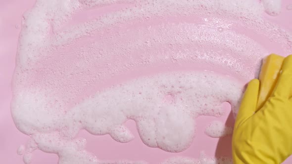 Hand in a Yellow Rubber Glove Holds a Cleaning Sponge and Wipes a Soapy Foam on a Pink Background