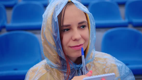 Young Woman in Raincoat with Notepad Pen Sitting on Stadium Bleachers Alone in Rainy Weather