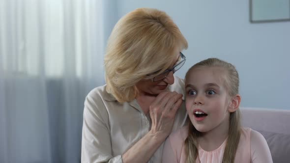 Positive Blond Grandmother Whispering Secret to Surprised Granddaughter, Family