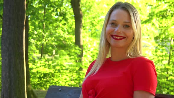 A Young Beautiful Woman Sits on a Bench in a Park on a Sunny Day and Smiles at the Camera - Closeup