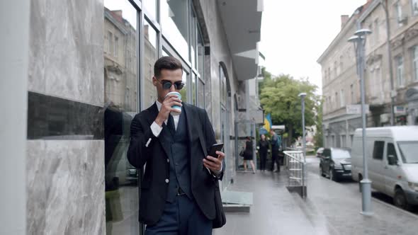 Man in Sunglasses Drinks a Cup of Beverage When Dials a Number on Phone Outdoors
