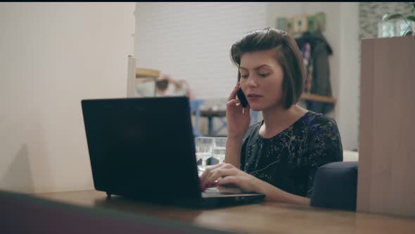 Young Business Woman Sitting By the Window with a Laptop and Talking on the Phone