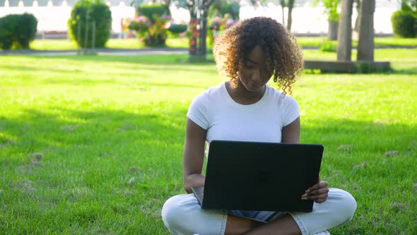 Young African American Woman Using Laptop Computer in Park. A Girl Works Remotely