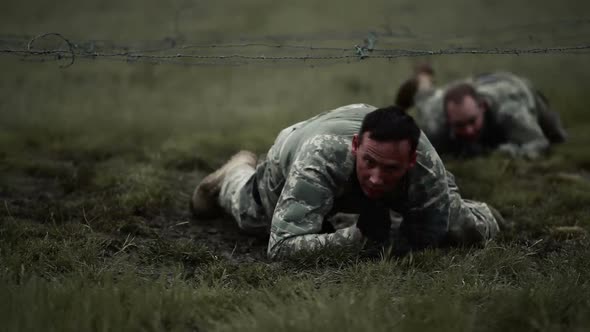 Soldiers crawling under low barbed wire at an obstacle course at a training, its muddy