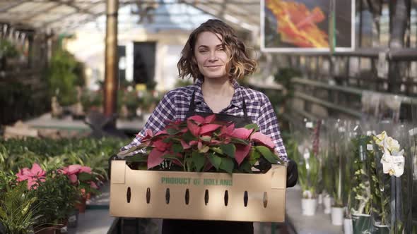 Beautiful European Smiling Female Gardener in Plaid Shirt and Black Apron Carrying Carton Box with