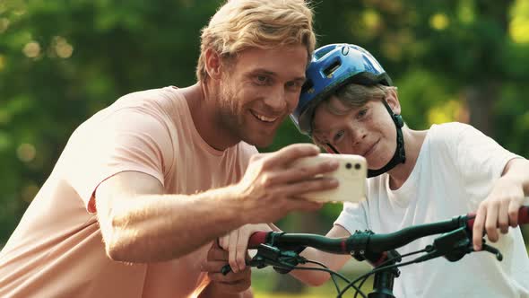 A smiling father is taking selfie photo with his son while he is teaching to ride a bicycle outdoors