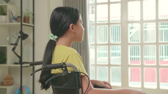 Asian Kid Girl Sitting In A Wheelchair Looking Out Of A Door