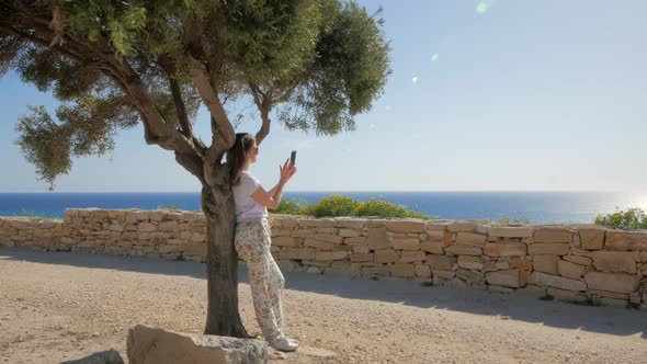 Woman Is Admiring Sea Views and Photographing By Mobile Phone, Leaning on Trunk of Tree