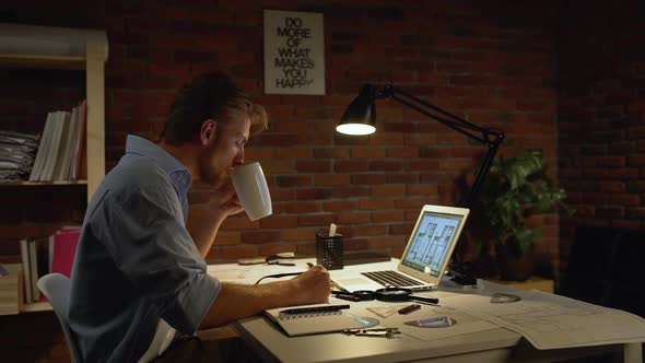 Caucasian Male with Light Beard and Blue Shirt Wearily Rinking From White Cup Near Computer at Table