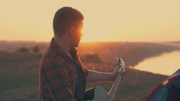 Guy Plays Guitar in Camp Against River and Setting Sun