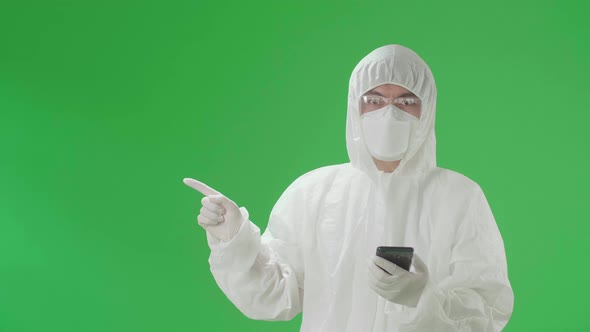 Asian Man Wear Protective Uniform PPE Use Mobile Phone With Pointing To Side In Green Screen Studio