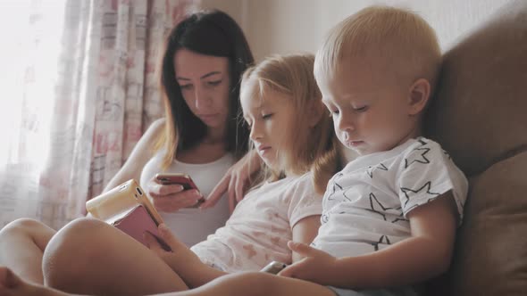 Young Mother, Daughter and Little Son Are Using Gadgets Lying on a White Bed. Concept Modern Family