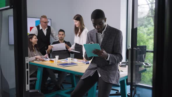 Thoughtful Absorbed African American Male Analyst Surfing Internet on Tablet As Colleagues