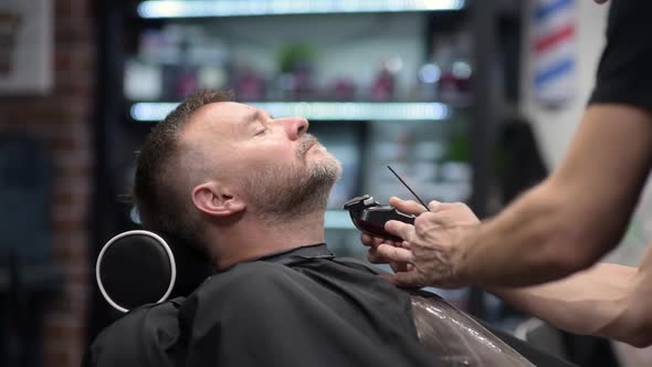 Barber master shaving handsome mature bearded man using electric shaver in salon