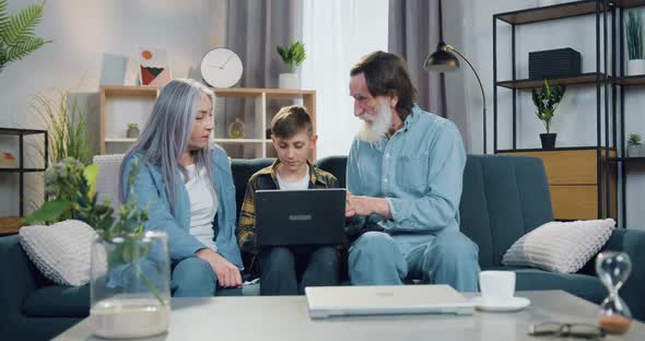 Boy Sitting on the Couch Between His Positive Old Grandparent and Working on Laptop