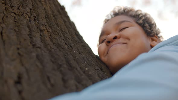 Close Up of Casual Cheerful Afro Female Embracing Tree with Closed Eyes in Park