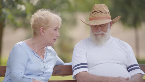 Elderly Caucasian Couple Sitting on the Bench in the Summer Park and Chatting. Positive Mature Man