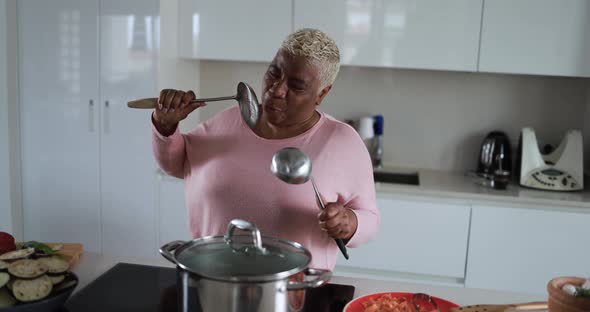 Happy black mature woman having fun cooking and dancing inside kitchen at home