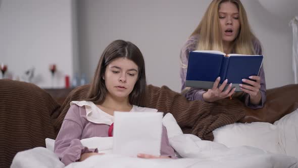 Absorbed Young Woman Sitting on Couch in Dorm Room As Blurred Friend Reading at Background