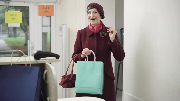 Portrait of Happy Elegant Senior Woman Standing in Shop with Shopping Bags and Waving at Camera