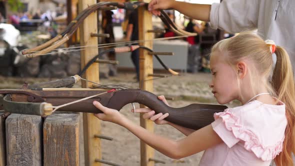 Blond Child Girl Aims and Shoots with Crossbow at Open Air Amusement Park Medium Shot