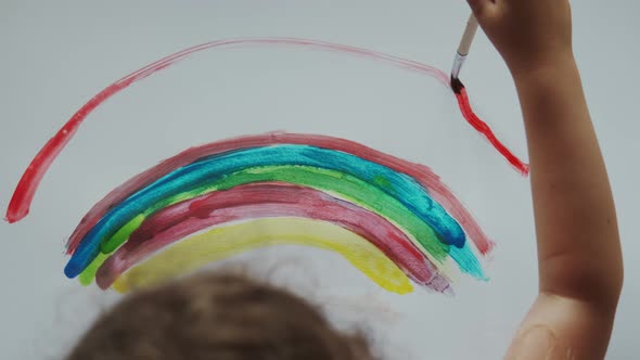 Little Girl Holds a Brush in Her Hand and Paints a Rainbow with Colored Paints on White Paper