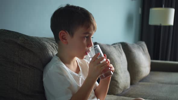 Child is Drinking a Glass of Water Sitting on Sofa at Home While Watching TV