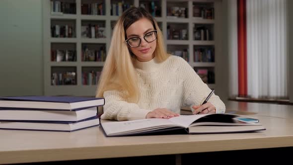 A Student Girl with Glasses Is Sitting at a Library Table with Books and Writing