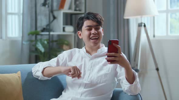 Smiling Asian Man Having Video Call On Smartphone While Lying On Sofa In The Living Room