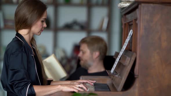 A Beautiful Woman Plays the Piano in Their Beautiful Apartment for Her Man Who Is Reading a Book