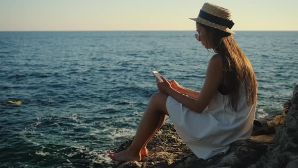 Woman is Typing on Smartphone Sitting on Cliff Over Sea in Summer