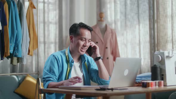 Asian Male Designer With Sewing Machine Talking On Smartphone While Designing Clothes On Laptop