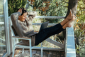 Middle-aged woman working on laptop computer sitting at table on wooden balcony of country house. - PhotoDune Item for Sale