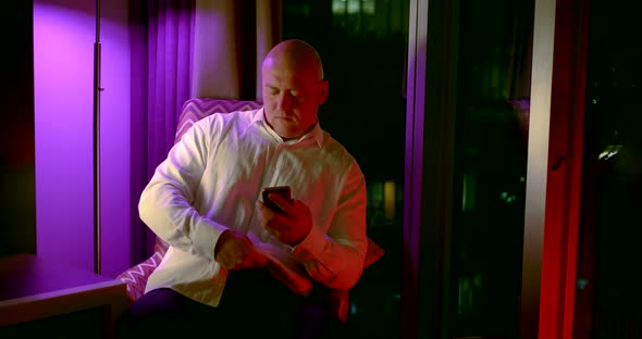 a Bald Man Sits on a Chair Near a Large Window. He Straightens His Shirt, Checks His Smartphone