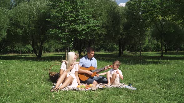 Family Picnic in Nature, Parents and Children Sit on a Blanket in a City Park, Father Plays Guitar