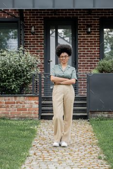 full length of smiling african american real estate agent standing with folded arms near cottage