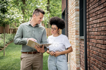 smiling real estate agent showing documents to african american woman near new city cottage