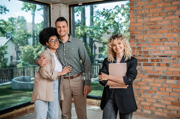 successful realtor with folder and cheerful multiethnic couple looking at camera in new cottage