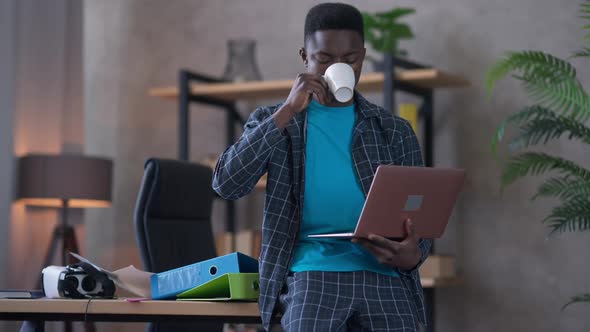 Confident Young African American Man Drinking Morning Coffee Sitting on Table Holding Laptop