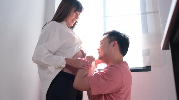 Asian man or husband kiss belly of his pregnant wife in front of glass windows with morning light