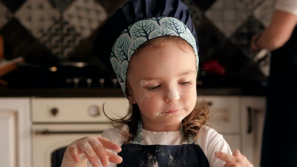 Cute Little Girl Smeared with Flour Licks Her Fingers with Dough in the Kitchen.