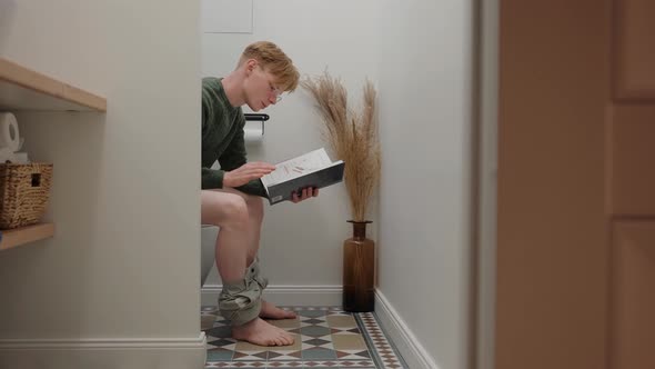 Young Man Reading a Book Sitting in White Modern Toilet