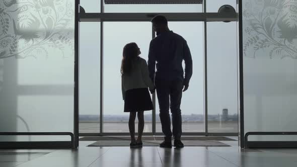 Back View Silhouettes of Adult Man and Little Girl Standing at the Airport Door and Looking at the