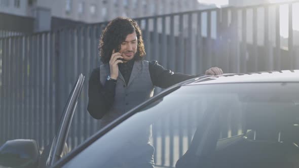 Positive Smiling Successful Elegant Businessman Standing at Open Car in Sunrays Talking on Phone