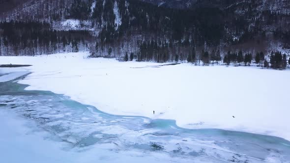 Two people are standing in a big snow field in a valley with a icey lake.